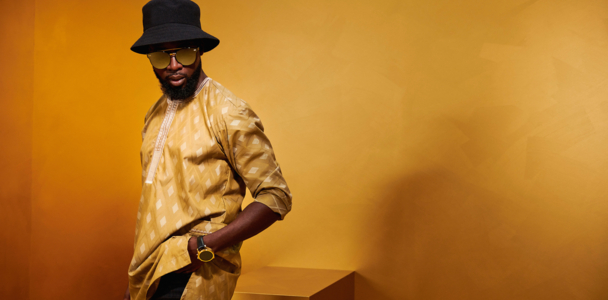 Un homme sûr de lui dans un Fokus doré de Getzner Textil, complété par un chapeau et des lunettes de soleil à la mode, incarne l'élégance africaine moderne.
